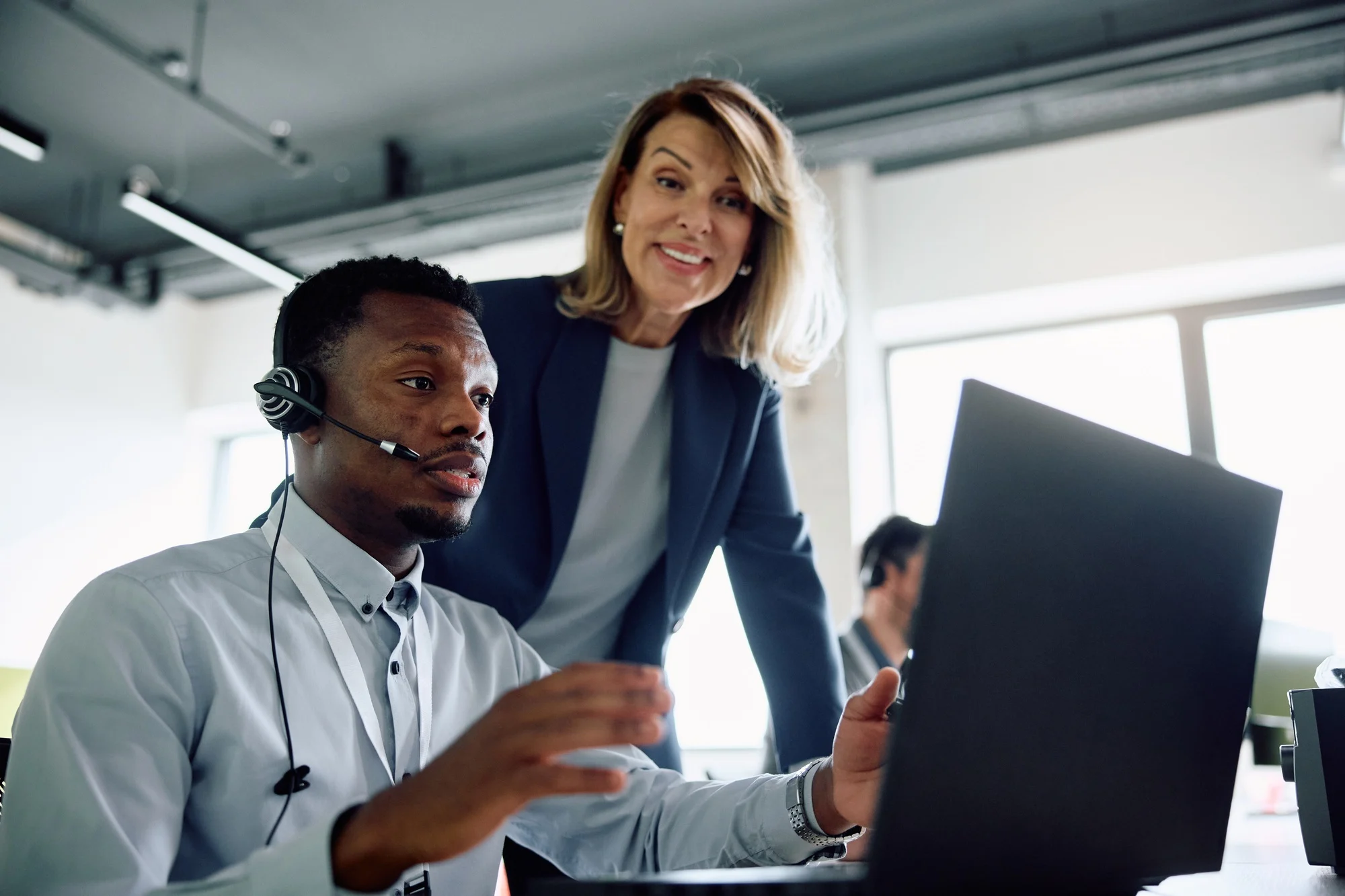 Black businessman with headset working on laptop while female CEO is supervising him in the office. Black businessman with headset working on laptop while female CEO is supervising him in the office.