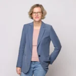 Eine Person mit kurzem, blondem Haar und Brille steht vor einem schlichten, hellen Hintergrund, trägt einen blauen Blazer über einem rosa Hemd und blaue Jeans, lächelt und hat eine Hand in der Tasche.