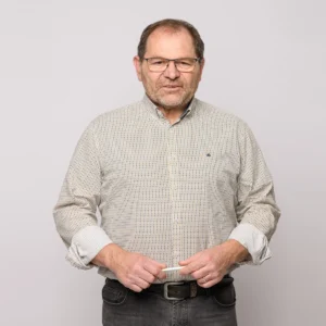 Ein Mann mittleren Alters mit Brille, kurzen Haaren und gestutztem Bart steht vor einem schlichten, hellen Hintergrund. Er trägt ein gemustertes Hemd und eine dunkle Jeans, wobei er seine Hände locker in der Nähe seiner Taille hält.