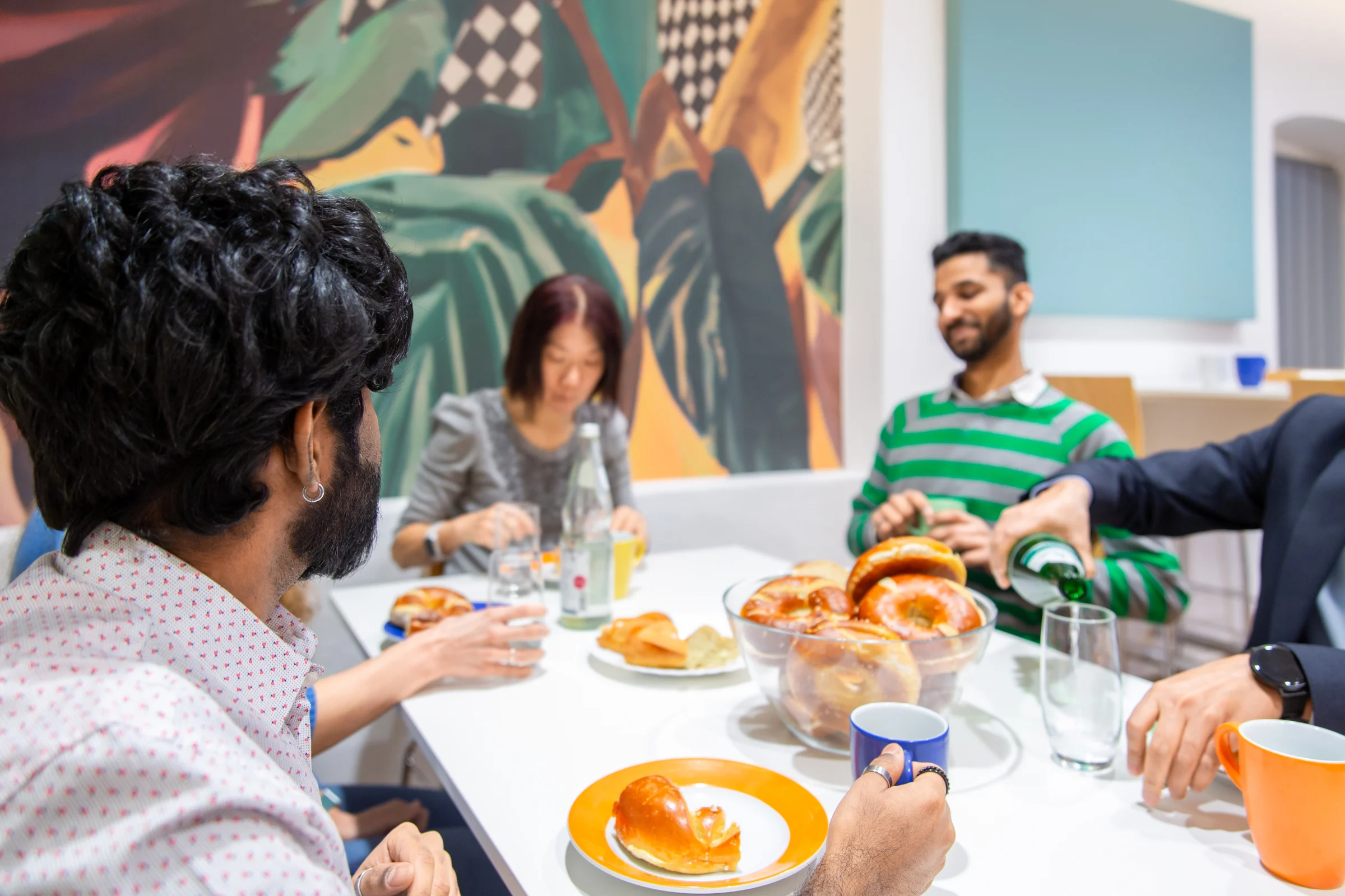 Vier Personen sitzen um einen Tisch und genießen Getränke und Gebäck. Eine Person gießt Wasser in ein Glas, während die anderen Tassen und Teller haben. An der Wand im Hintergrund ist eine bunte Wandmalerei zu sehen.