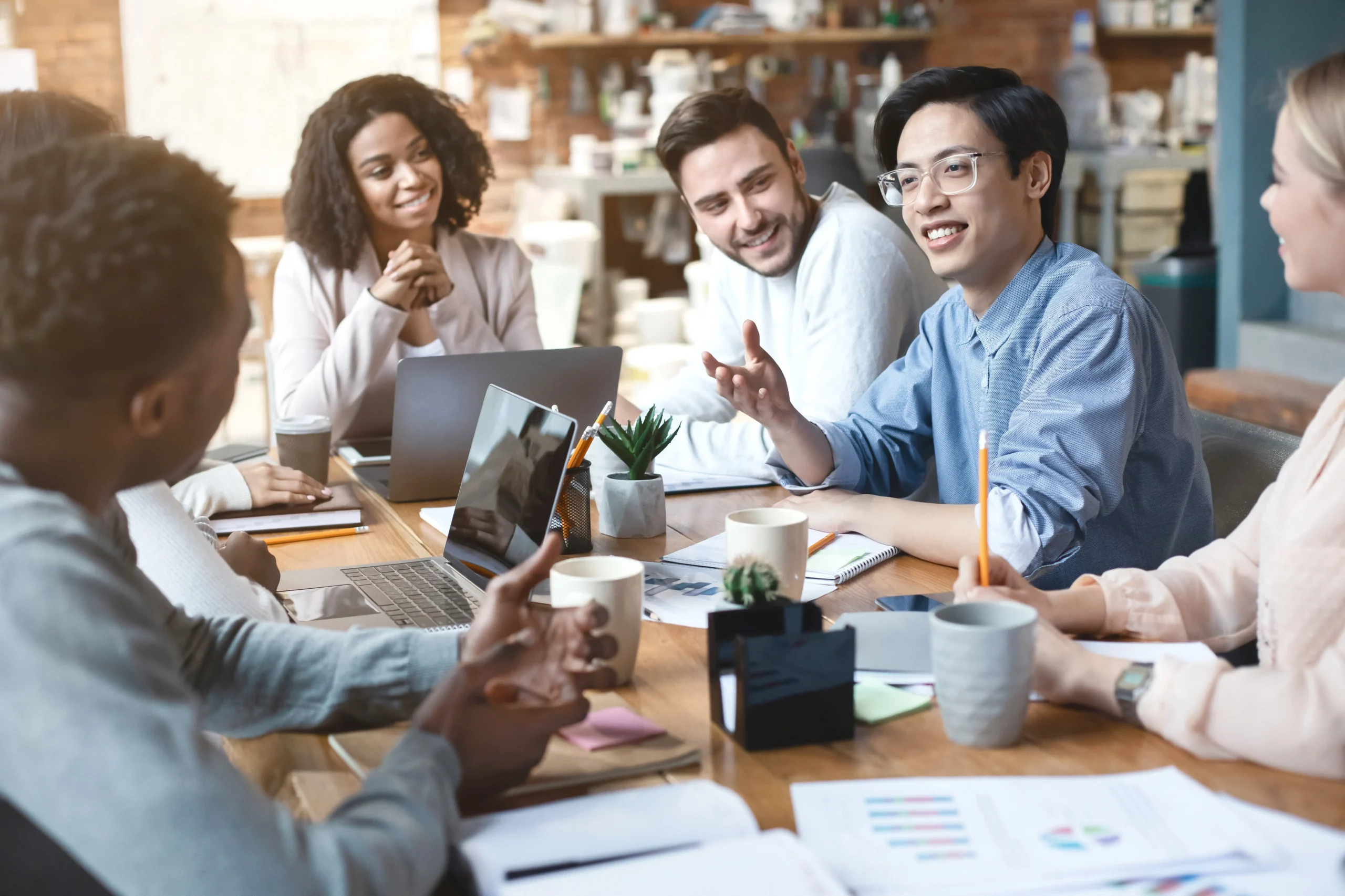 1-6 Eine bunt gemischte Gruppe von sechs Personen sitzt um einen Tisch in einem Büro und führt eine lebhafte Diskussion mit Laptops, Papieren und Kaffeetassen vor sich. Sie lächeln und scheinen gemeinsam an einem Projekt zu arbeiten.