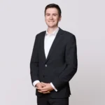 Ein junger Mann in einem dunklen Anzug steht lächelnd mit gefalteten Händen vor einem schlichten Hintergrund.